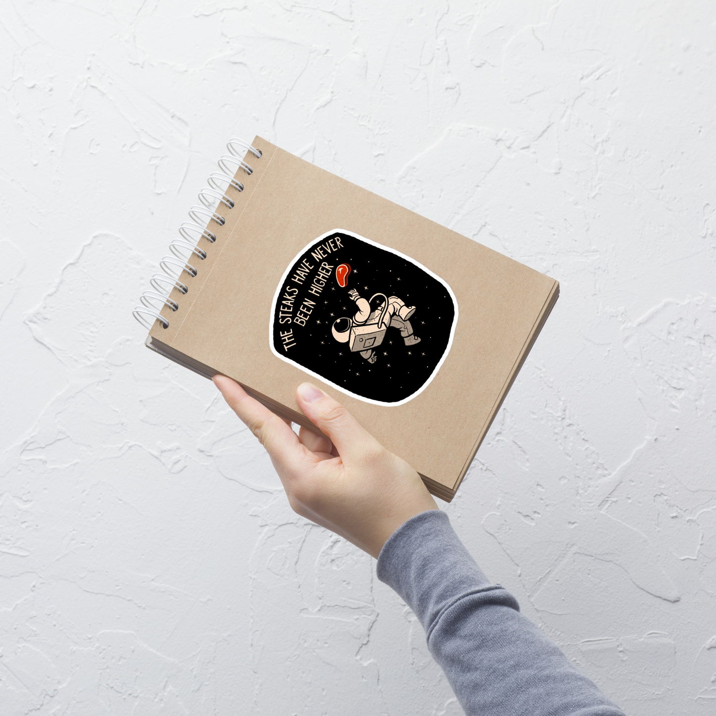 An astronaut floating through space with a small steak in front of him, paired with the phrase ‘The Steaks Have Never Been Higher’. A playful and humorous design that blends space exploration with a clever visual pun. Perfect for fans of witty wordplay and quirky art.