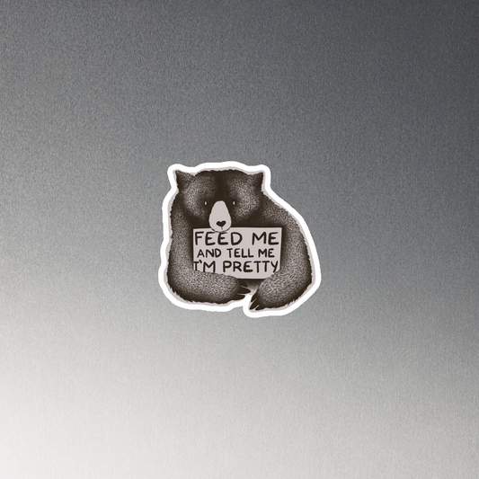 This drawing is of a cute, cartoon-style bear holding a sign with a heart-shaped nose and simple, expressive eyes. The sign reads, 'Feed me and tell me I'm pretty' in a handwritten, playful font. The bear's fur is detailed with fine, textured lines, giving it a cozy, endearing look. The drawing has a monochrome, black-and-white color scheme