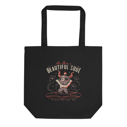 This design features a cute, cartoonish character with horns and a pentagram on its forehead, standing on a red circle with ghost-like figures surrounding it. Above the character, the text reads 'You have a beautiful soul,' and below, 'Give it to me.' The design contrasts dark and cute imagery with playful lettering.