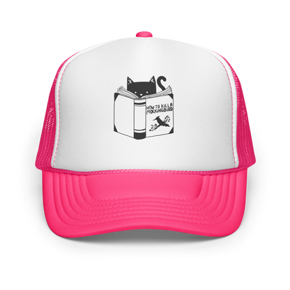 A black and white illustration features a cat peeking over an open book titled 'How to Kill a Mockingbird,' playfully altering the classic novel's title. The cat has a heart-shaped nose and simple, cute facial features. The design is whimsical and ironic, combining classic literature with a humorous twist.