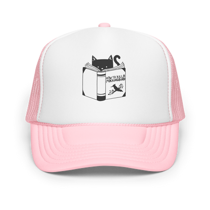 A black and white illustration features a cat peeking over an open book titled 'How to Kill a Mockingbird,' playfully altering the classic novel's title. The cat has a heart-shaped nose and simple, cute facial features. The design is whimsical and ironic, combining classic literature with a humorous twist.