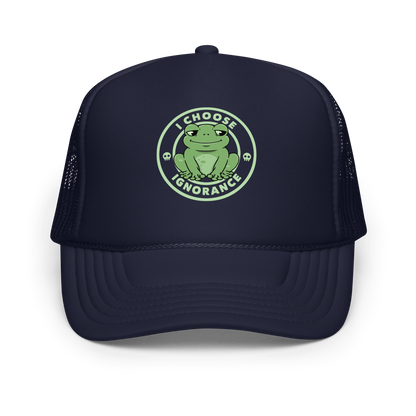The image shows a cartoonish green frog with a sly smile, sitting with legs splayed out. Around it, the phrase 'I CHOOSE IGNORANCE' is written in bold letters. Two small skull designs are included, one on each side of the frog. The style is playful and ironic, blending elements of humor and rebellion.