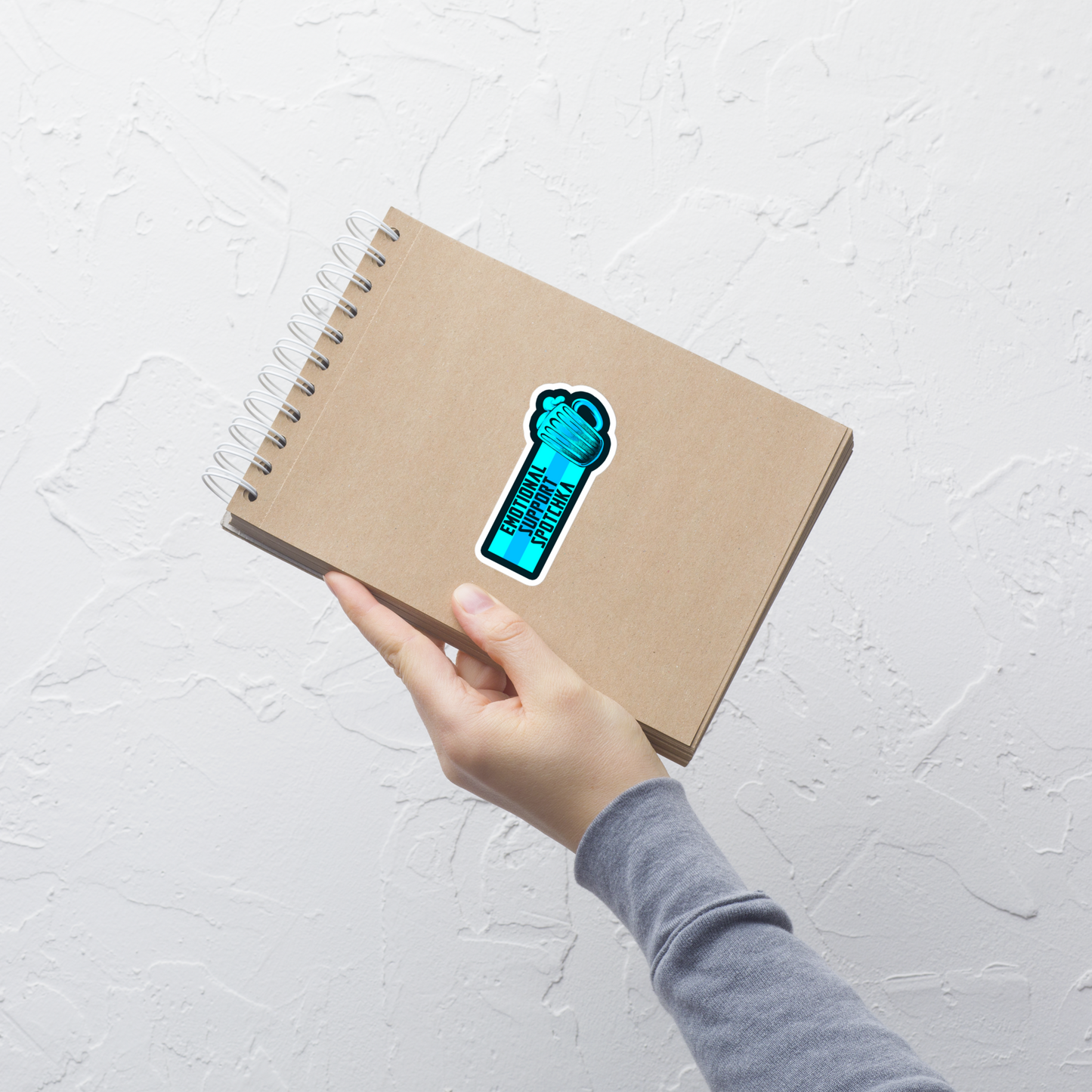 The design features a stylized mug filled with a frothy beverage, colored in vibrant shades of blue. Next to the mug, bold text reads 'EMOTIONAL SUPPORT SPOTCHKA'. The use of color and font style gives it a playful, pop-culture vibe, hinting at something fantastical or sci-fi influenced.
