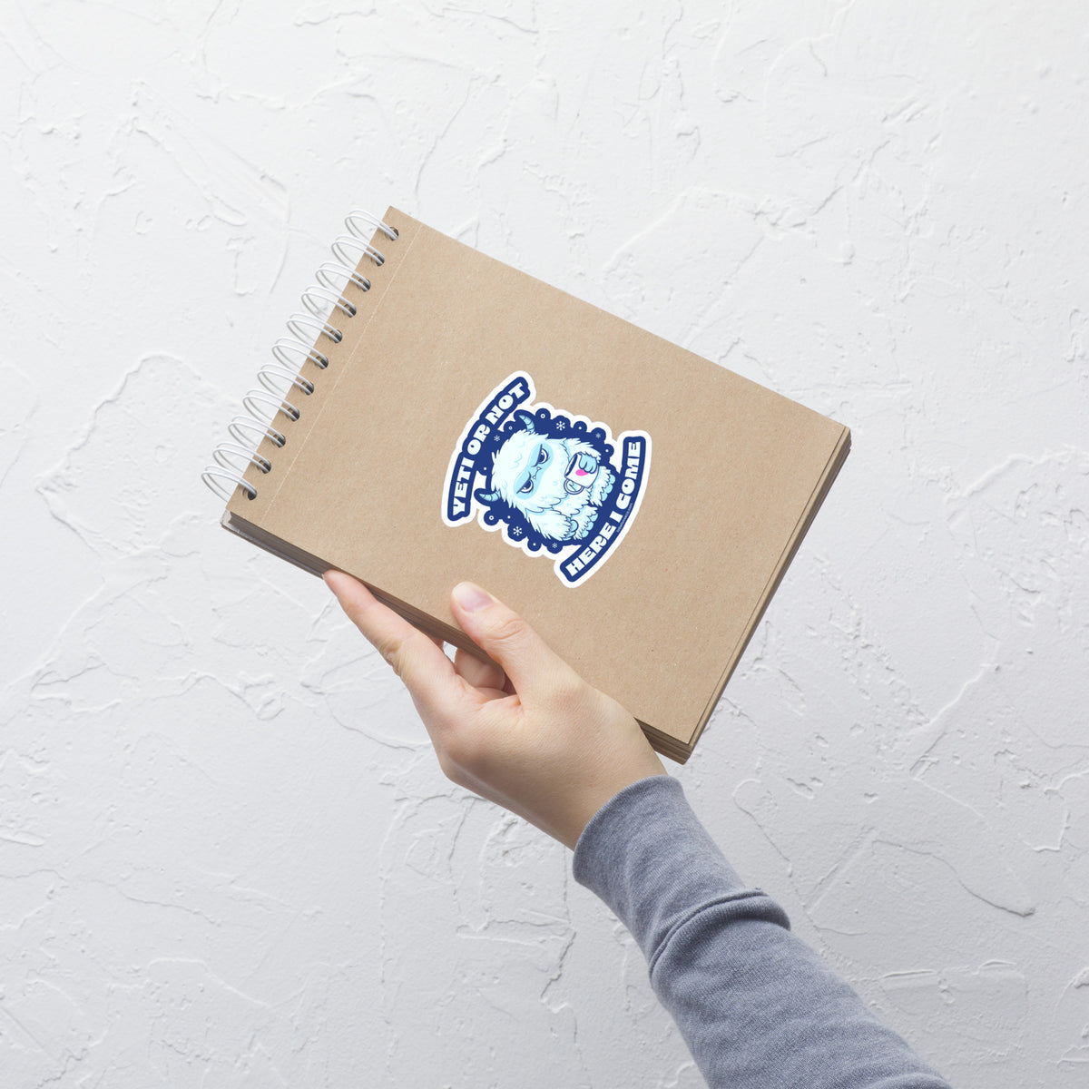 An illustration in shades of blue featuring an adorable yet grumpy little monster holding a cup of coffee, with the phrase "Yeti Or Not Here I Come". This humorous design plays on the classic phrase “Ready or not, here I come”, replacing “ready” with “yeti” for a playful and quirky twist.