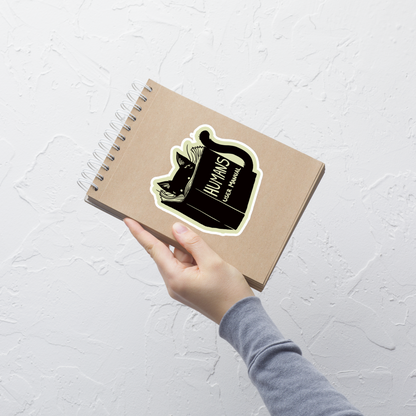 A kitten peeking out from behind a huge book titled "Humans: User Manual". This design plays on the idea that cats are trying to figure out how to control us. Perfect for cat lovers, book fans, and anyone who enjoys lighthearted humor in prints.