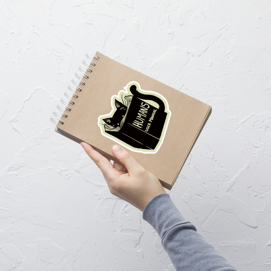 A kitten peeking out from behind a huge book titled "Humans: User Manual". This design plays on the idea that cats are trying to figure out how to control us. Perfect for cat lovers, book fans, and anyone who enjoys lighthearted humor in prints.