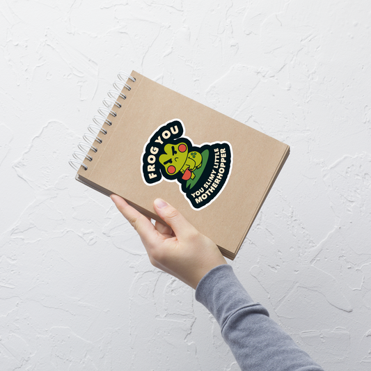 The design features a cartoonish green frog with exaggerated angry features, holding a teacup. The frog has round red cheeks. Above it, text says 'Frog You' and below, 'You Slimy Little Motherhopper'. The style is playful and humorous, reminiscent of cute comic art.