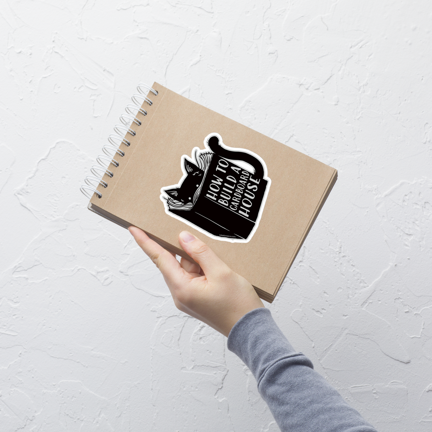 A minimalist illustration of a curious cat reading a book titled "How To Build A Cardboard House". A playful design that captures every cat’s true passion for cardboard boxes, perfect for cat lovers who appreciate subtle humor and clean, modern art.