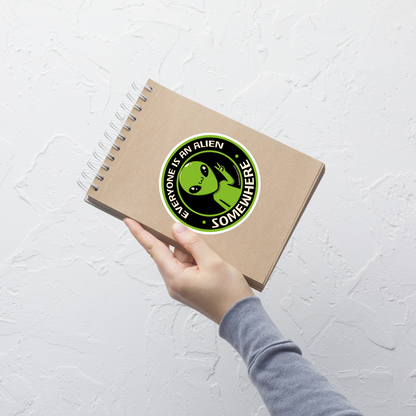A stamp-style design of a green alien making a “V” sign, paired with the phrase “Everyone Is An Alien Somewhere”. A playful artwork about feeling different and embracing individuality.