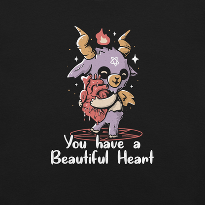 The design features a cute, cartoonish, anthropomorphic goat with big horns and a flame above its head, standing on a red pentagram. The goat has a star on its forehead and is hugging a large, anatomically correct human heart. The text below reads, 'You have a Beautiful Heart'. The image combines elements of occult symbolism with a playful, endearing aesthetic.