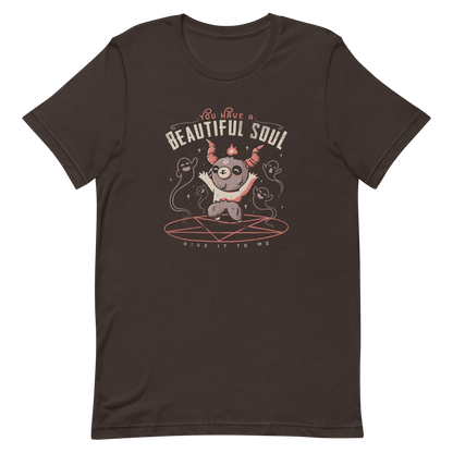 This design features a cute, cartoonish character with horns and a pentagram on its forehead, standing on a red circle with ghost-like figures surrounding it. Above the character, the text reads 'You have a beautiful soul,' and below, 'Give it to me.' The design contrasts dark and cute imagery with playful lettering.