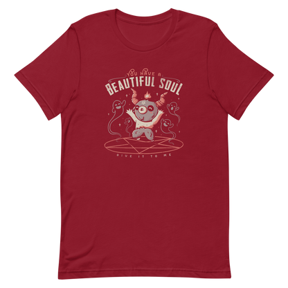 This design features a cute, cartoonish character with horns and a pentagram on its forehead, standing on a red circle with ghost-like figures surrounding it. Above the character, the text reads 'You have a beautiful soul,' and below, 'Give it to me.' The design contrasts dark and cute imagery with playful lettering.