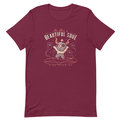This design features a cute, cartoonish character with horns and a pentagram on its forehead, standing on a red circle with ghost-like figures surrounding it. Above the character, the text reads 'You have a beautiful soul,' and below, 'Give it to me.' The design contrasts dark and cute imagery with playful lettering.